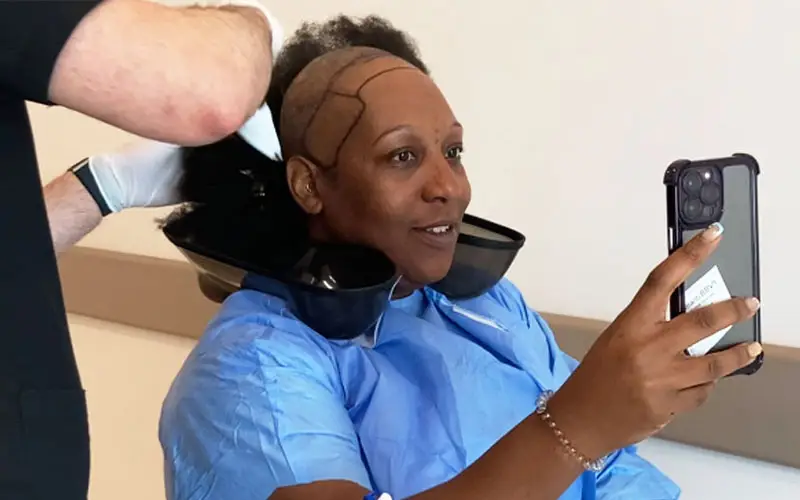 An Afro hair transplant patient holds her phone to record a video of herself while her hair is being shaved.